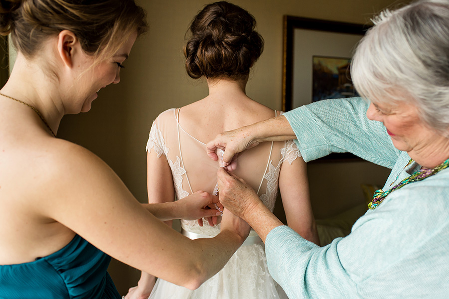 Maid of honor and mother of the bride helping bride into her wedding gown.