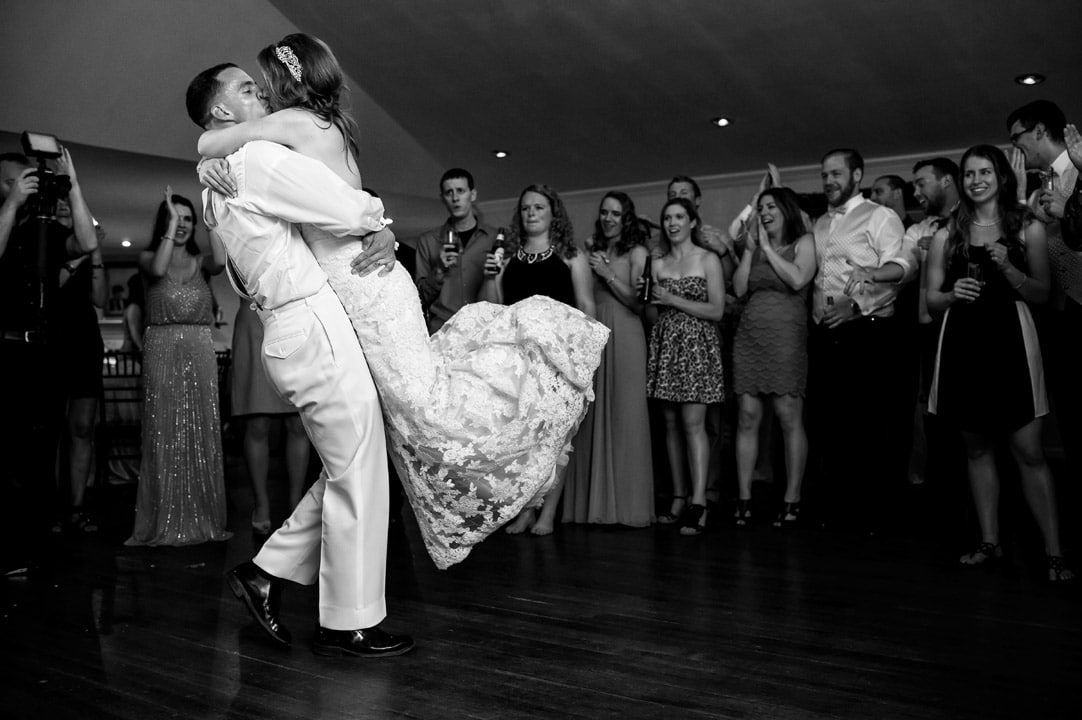 Groom lifts bride and carries her across dance floor at wedding reception.