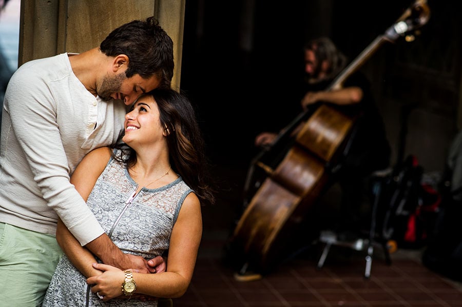 An engaged couple hug as a street performer plays Cello behind them during their engagement session in Central Park.