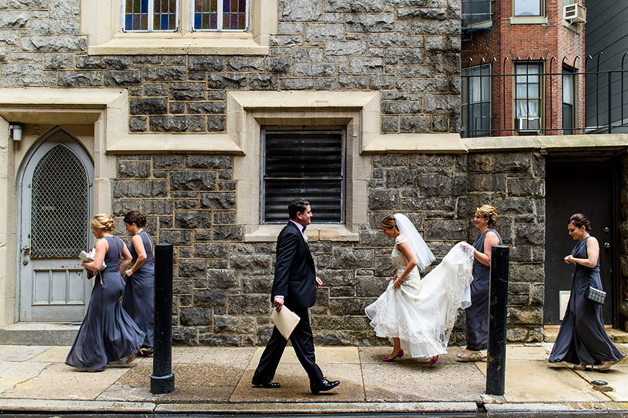 Bride and bridesmaids walk down rainy alley to church ceremony on wedding day.