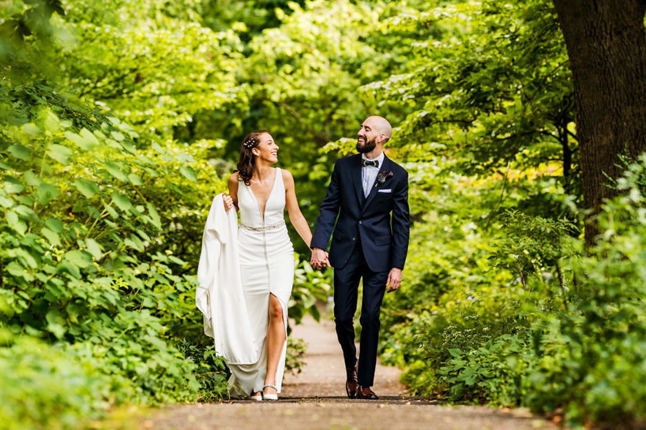 Bride and groom hold hands while smiling on a path through lush green foliage.