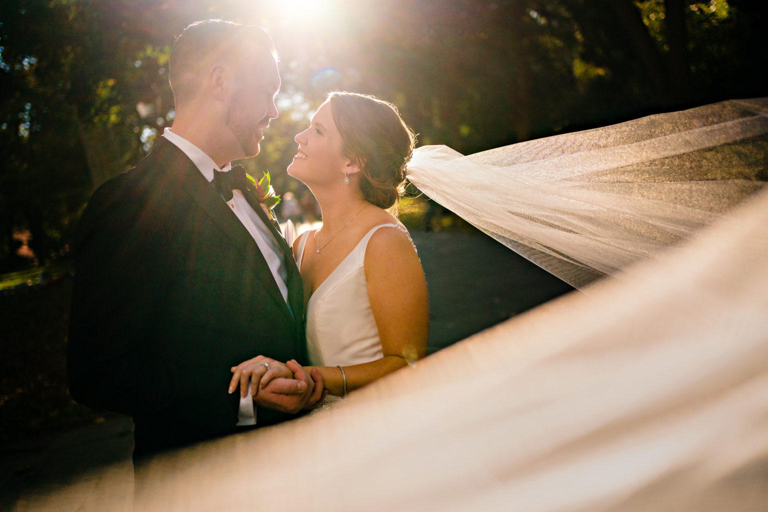 Bride and groom hug each other with warm sun flare behind them while bride's cathedral length veil flys around them.